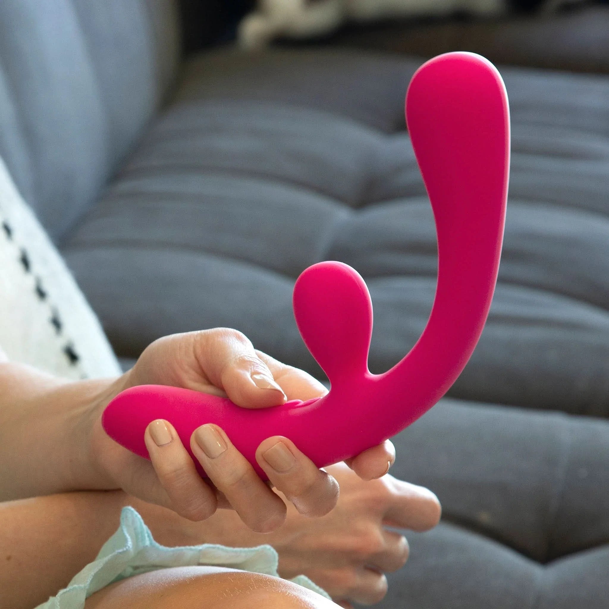 A person holding the JIMMYJANE® Reflexx™ 3, a pink curved silicone vibrator designed for g-spot stimulation, while sitting on a gray couch.