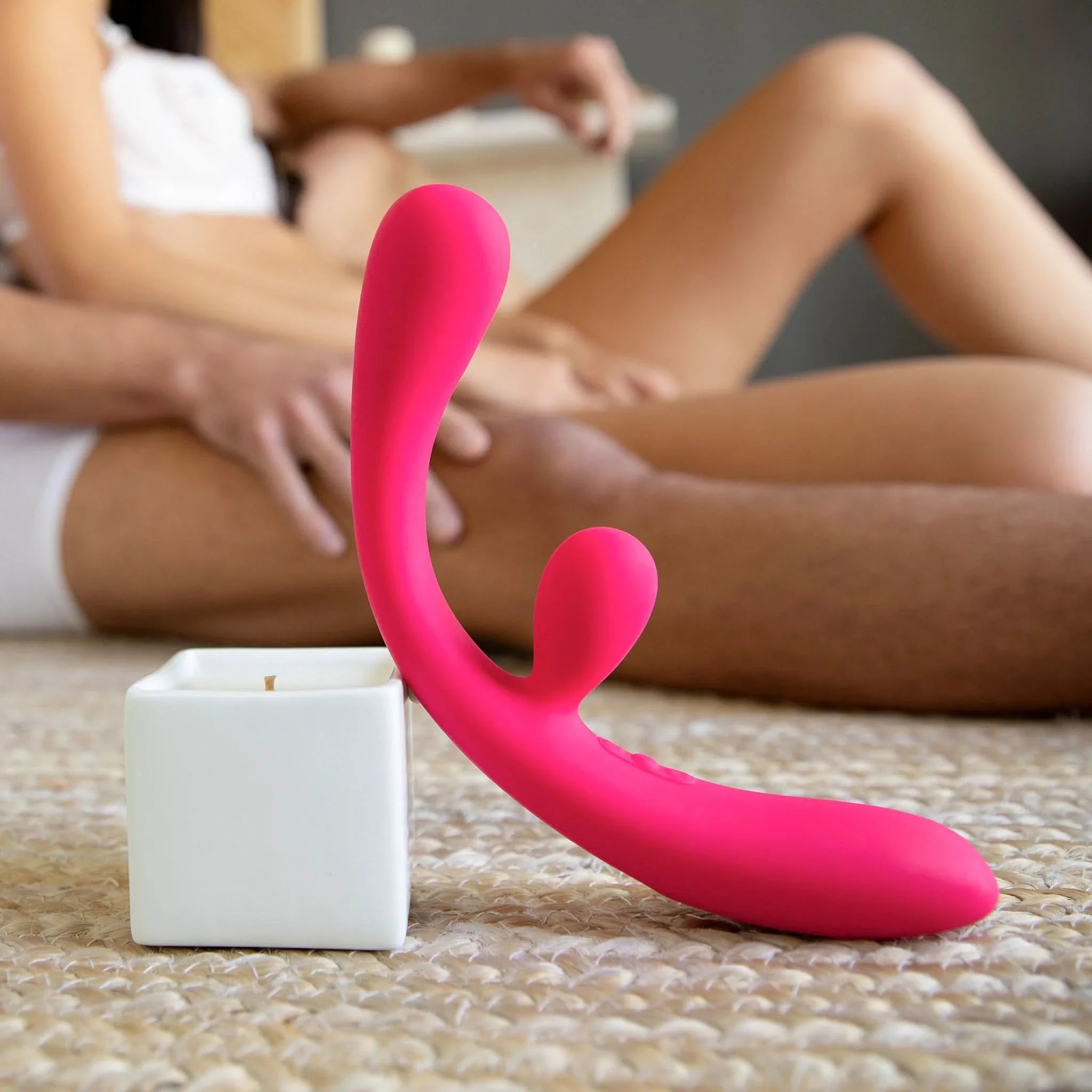 The JIMMYJANE® Reflexx™ 3 pink silicone vibrator with dual motors rests on a textured rug as a couple sits closely together in the background.