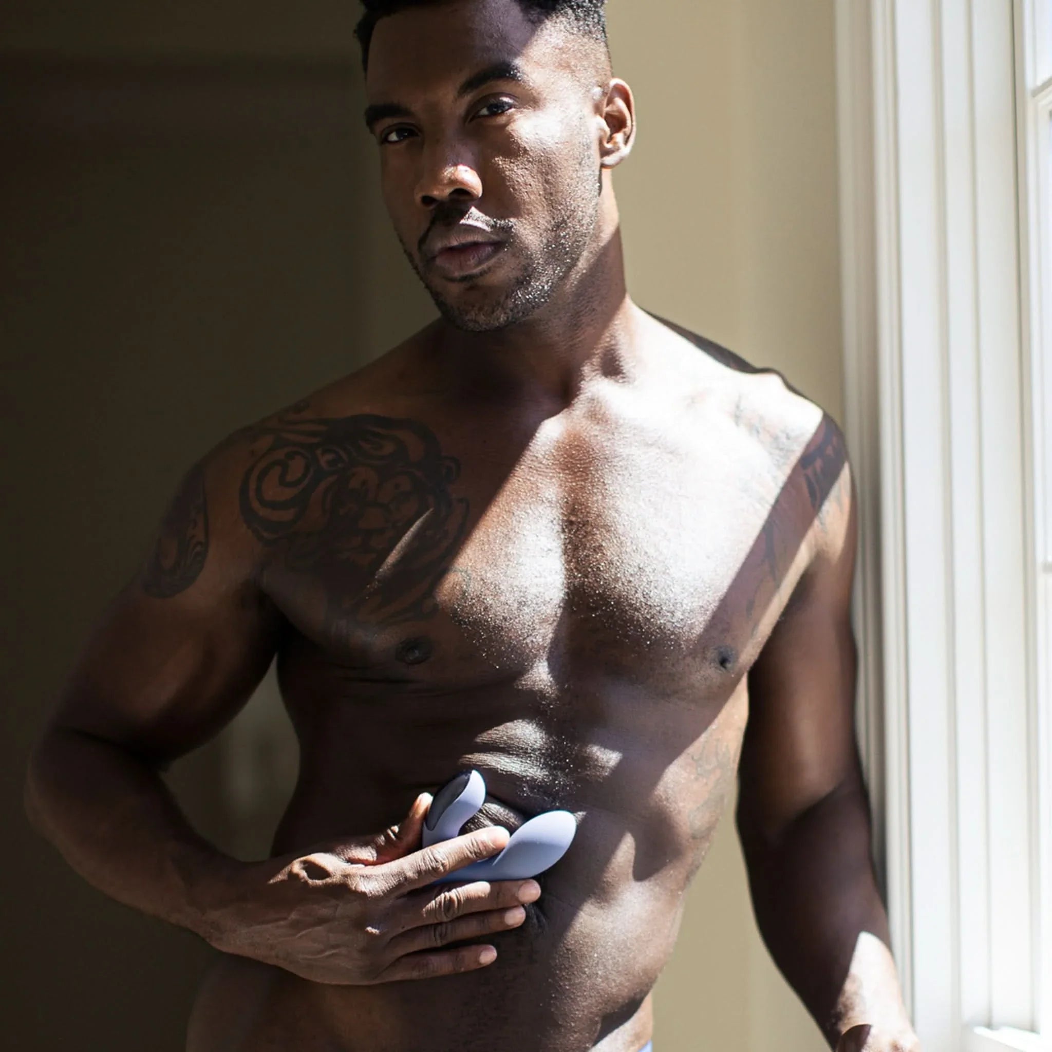 A shirtless, tattooed man stands by a sunlit window, holding the JIMMYJANE® Neptune 2™ with Remote prostate massager against his torso.