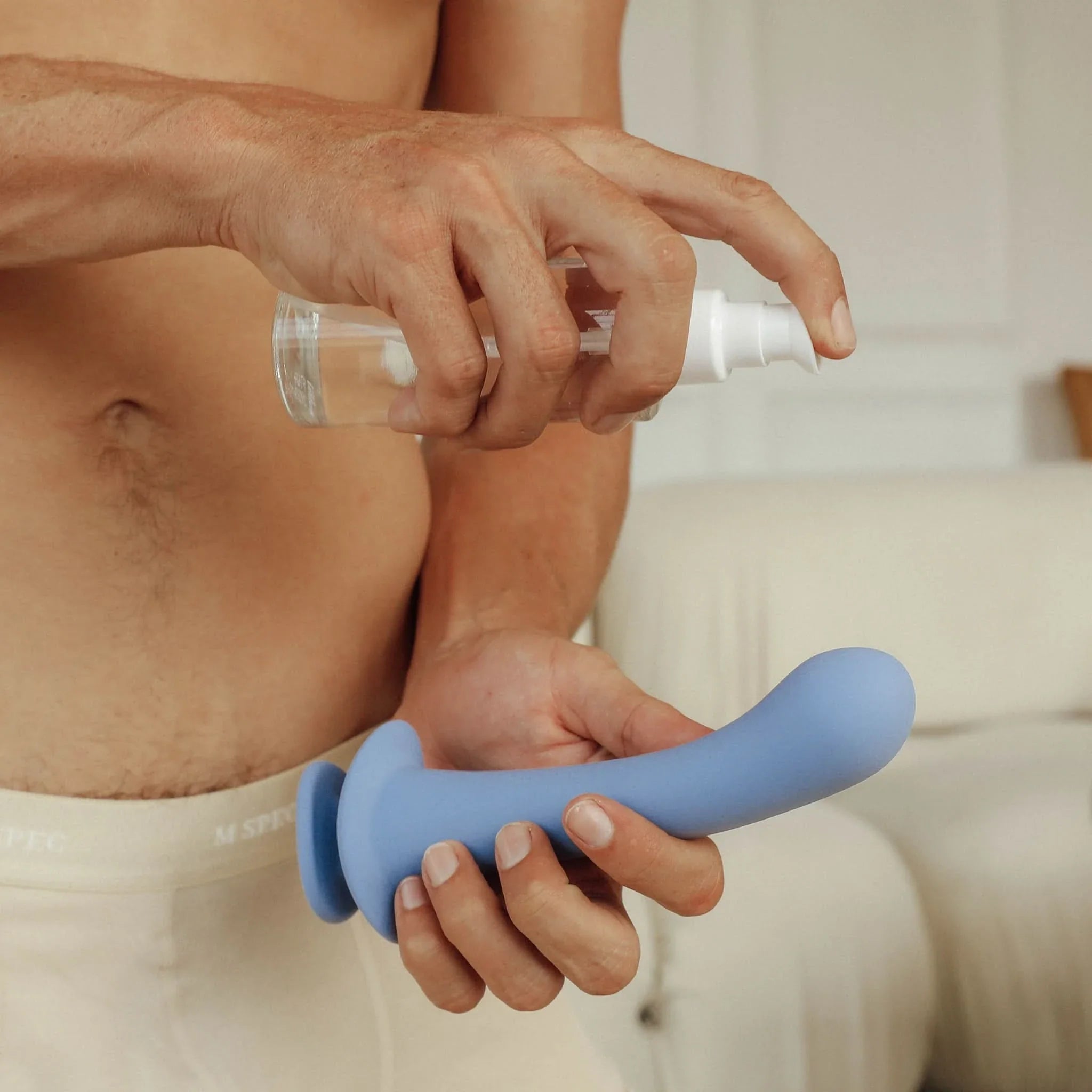 A person sprays liquid onto a blue JIMMYJANE® Cyra™ 6-inch silicone dildo indoors while wearing light underwear.