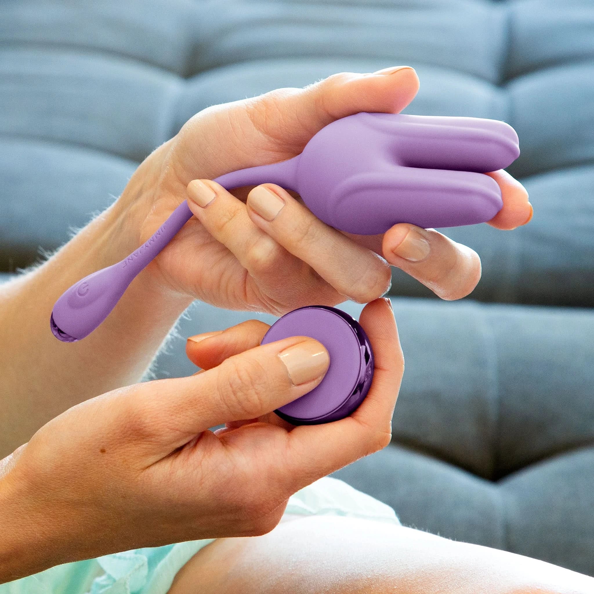 A person holds a purple JIMMYJANE® Form 2® Kegel Trainer and remote while sitting on a gray couch, ready to strengthen their pelvic floor muscles.