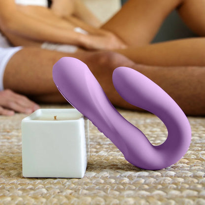 A JIMMYJANE® Reflexx™ 1 vibrator with dual motors and a white candle rest on a woven rug, while two people sit blurred in the background.