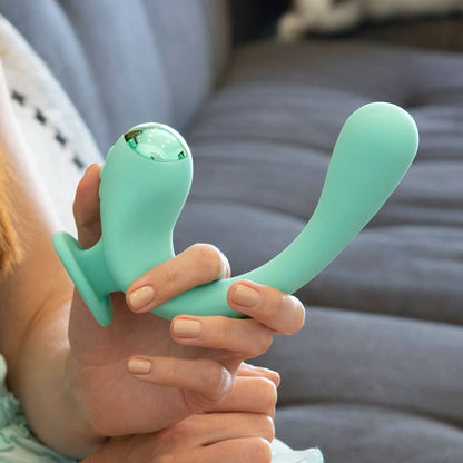 A hand holds the teal JIMMYJANE® Reflexx™ Gripp™ 2, a curved silicone vibrator with a flared base for precise G-spot or clitoral stimulation, against a gray couch background.