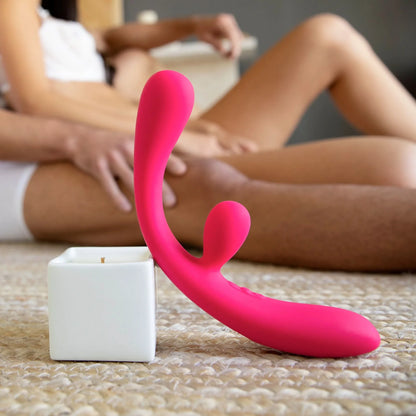 The JIMMYJANE® Reflexx™ 3 pink silicone vibrator with dual motors rests on a textured rug as a couple sits closely together in the background.