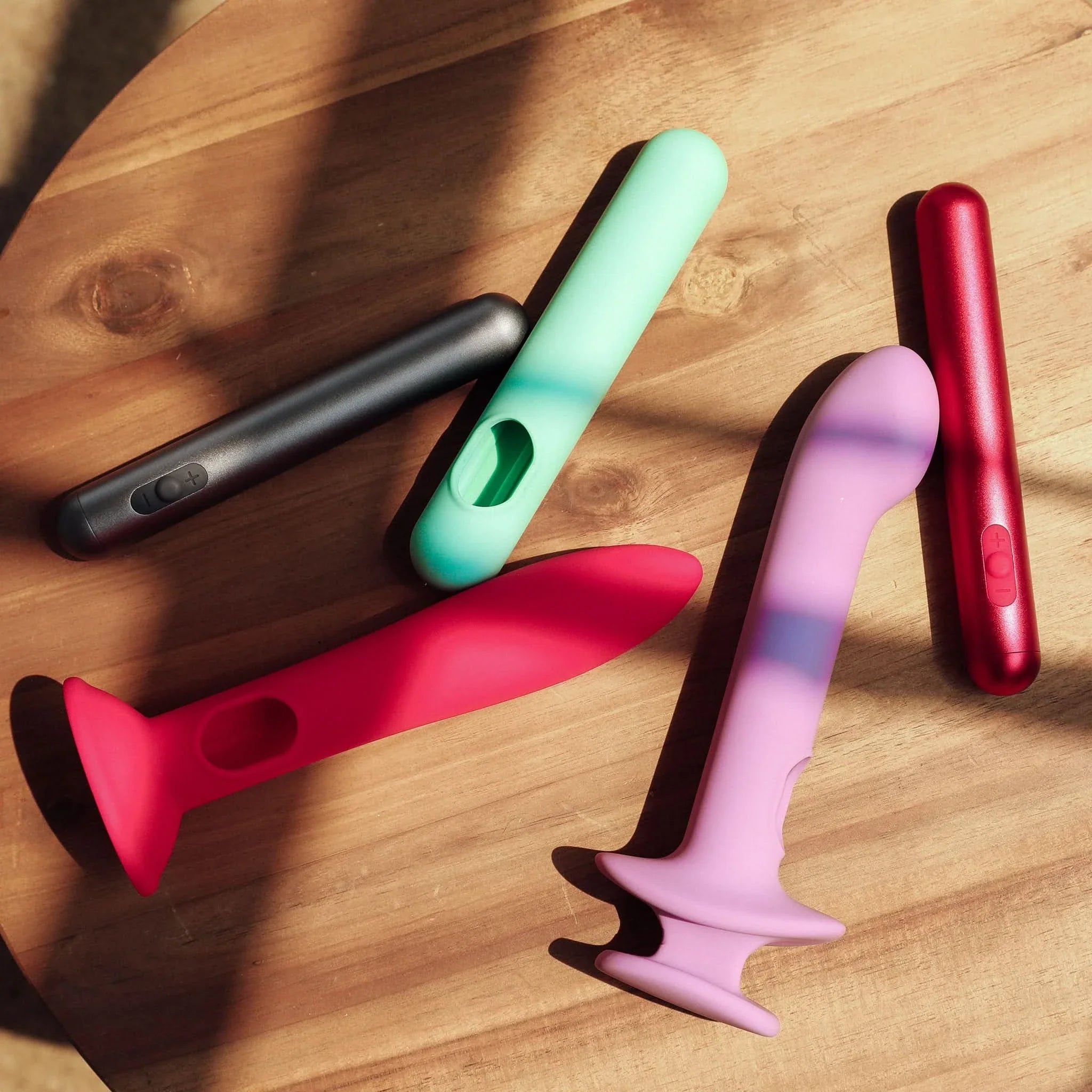 Four vibrant silicone and metal JIMMYJANE® vibrators, including the waterproof bullet and sleek Chroma™, are displayed on a wooden surface under sunlight.