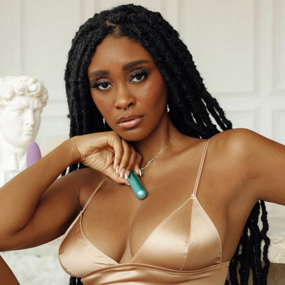 A woman with long braids, wearing a beige satin top, sits indoors near a statue while holding the JIMMYJANE® Chroma™ Mini with Remote, a teal mini vibrator.