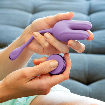 A person holds a purple JIMMYJANE® Form 2® Kegel Trainer and remote while sitting on a gray couch, ready to strengthen their pelvic floor muscles.