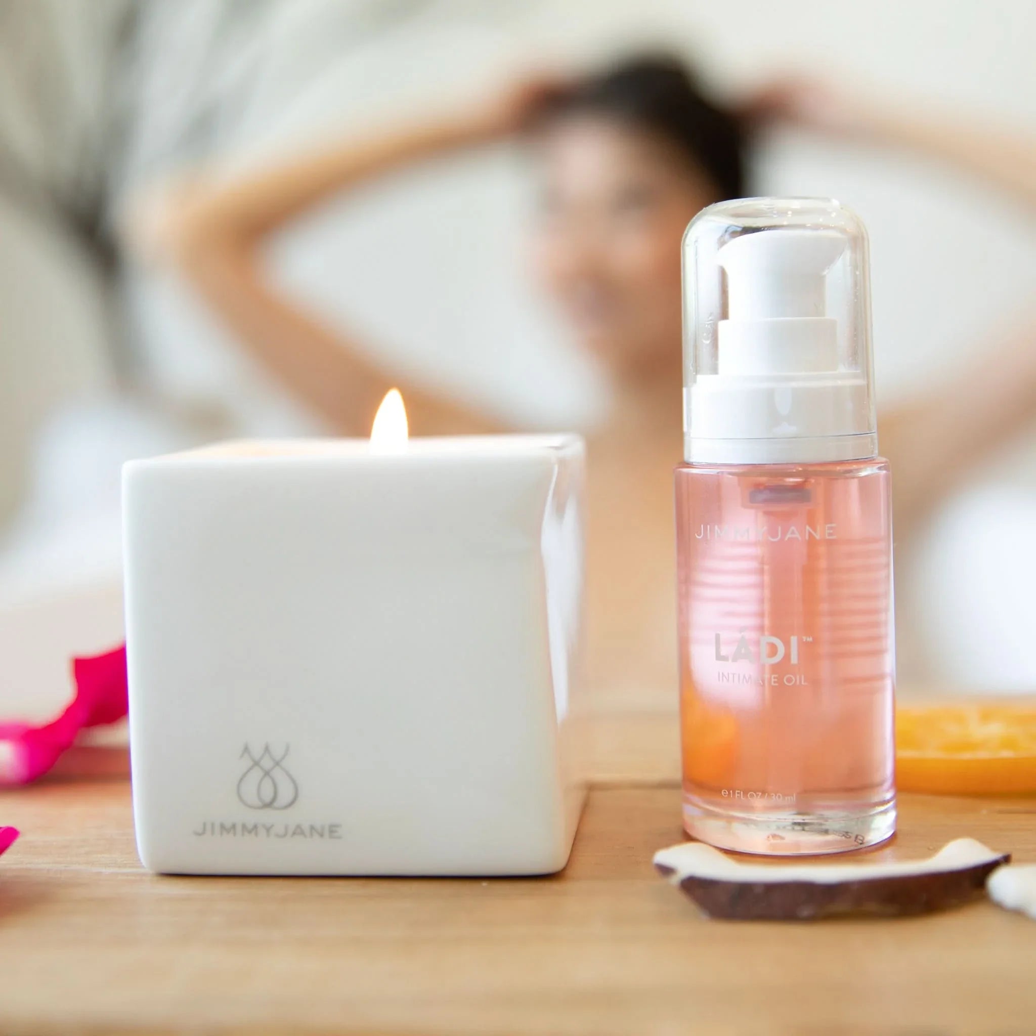 A lit candle and a bottle of JIMMYJANE® Ládi™ Oil, known for relieving vaginal dryness, sit on a wooden surface with a blurred person in the background.