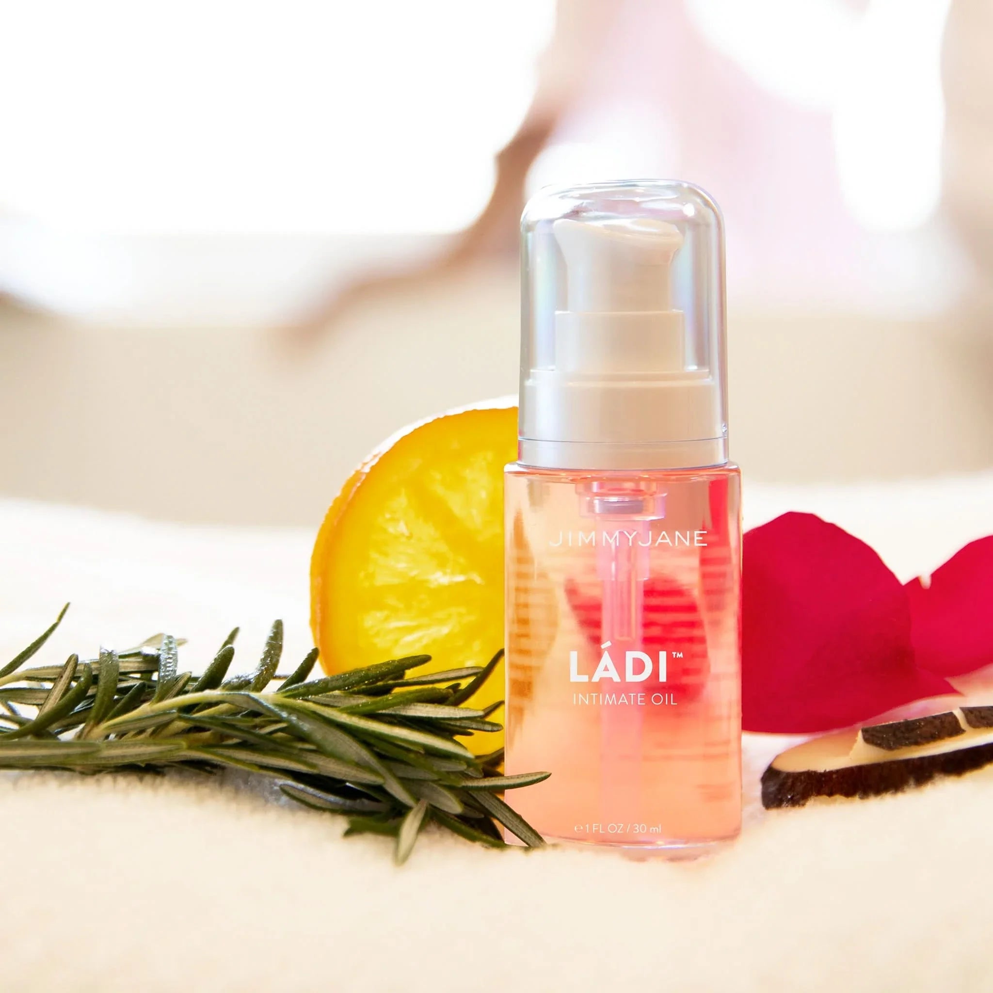 A bottle of JIMMYJANE® Ládi™ Oil, surrounded by rosemary, orange slice, rose petals, and wood—ideal for skin hydration and vaginal dryness relief.