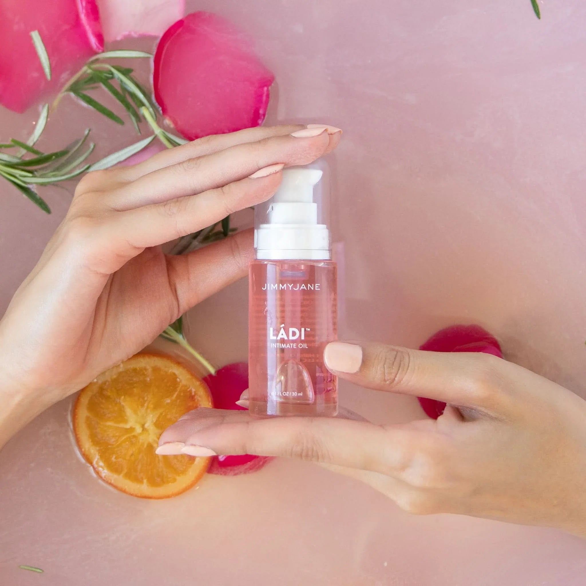 Hands holding a bottle of Ládi™ Oil by JIMMYJANE® with rose petals, rosemary, and orange slice on a pink surface—ideal for hydrating skin and relieving vaginal dryness.