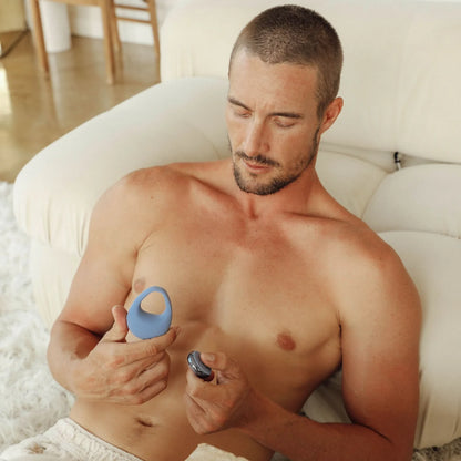 A shirtless man sits on a couch holding the JIMMYJANE® Kore™ with Remote, a blue vibrating silicone C-ring, and its small remote control in his hands.