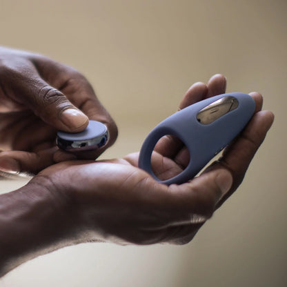 Close-up of hands holding the blue JIMMYJANE® Tarvos™ with Remote vibrating silicone c-ring and its round charger, highlighting the sleek design for hands-free clitoral stimulation.