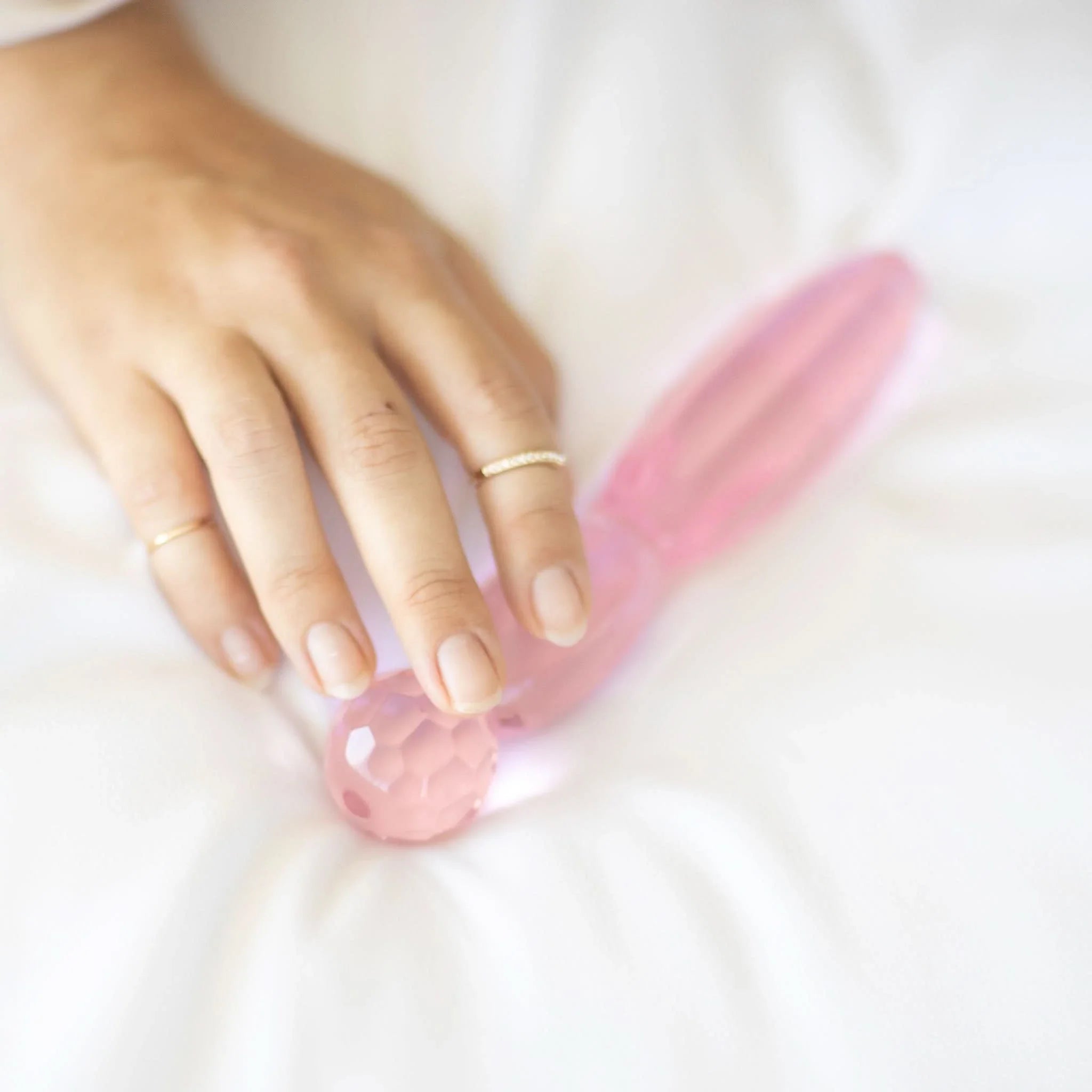 A hand adorned with gold rings rests on the JIMMYJANE® Cissus 7-inch, a pink, faceted borosilicate glass object placed on a white fabric surface.