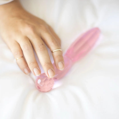 A hand adorned with gold rings rests on the JIMMYJANE® Cissus 7-inch, a pink, faceted borosilicate glass object placed on a white fabric surface.